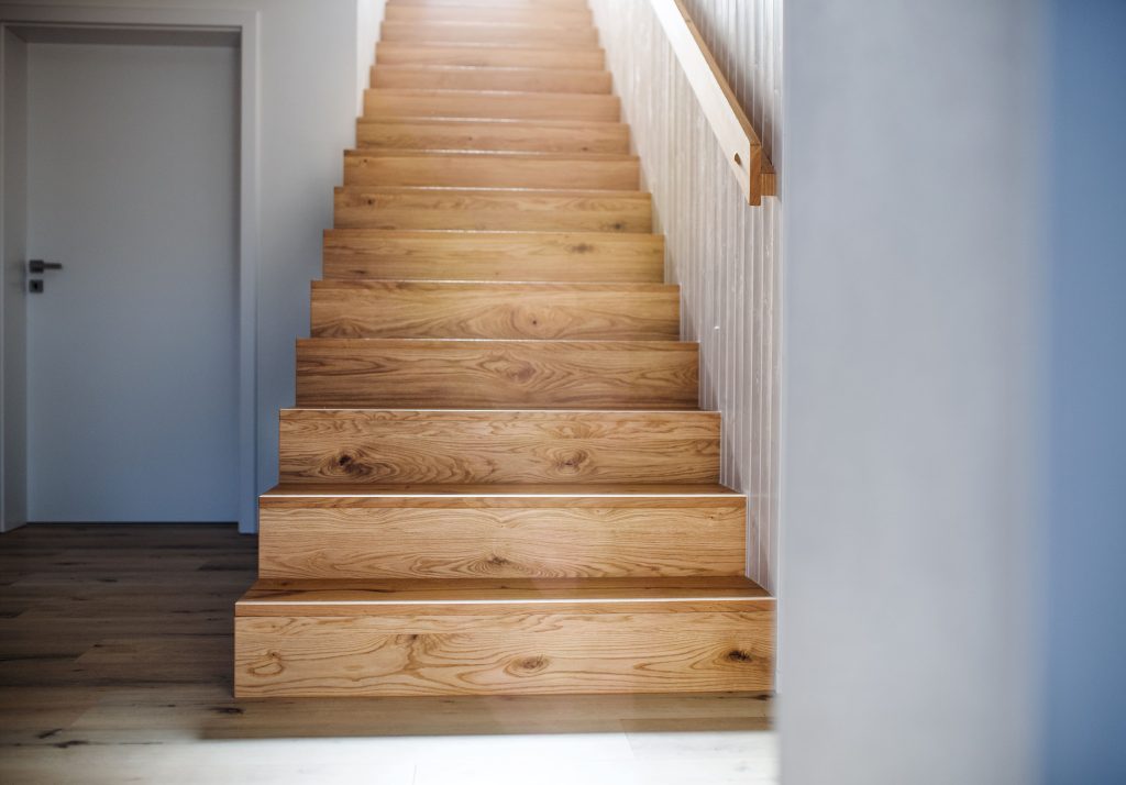 A flight of light wooden stairs rises beside a white paneled wall, with a closed gray door to the left.