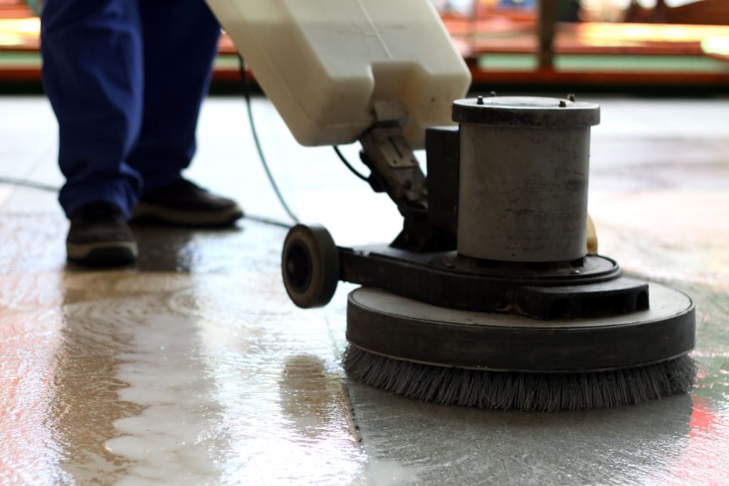 A person in blue work pants operates a rotary floor scrubber with circular bristles on a wet floor.