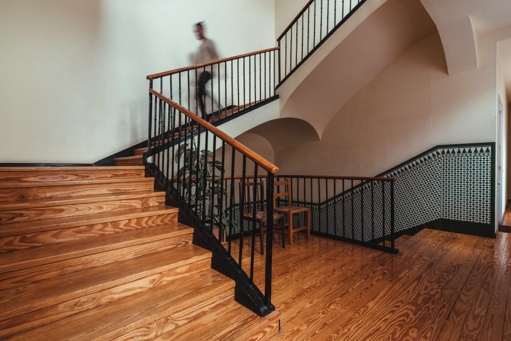 A person walks on a multi-level wooden staircase with black metal railings in an interior featuring arches and patterned tilework.