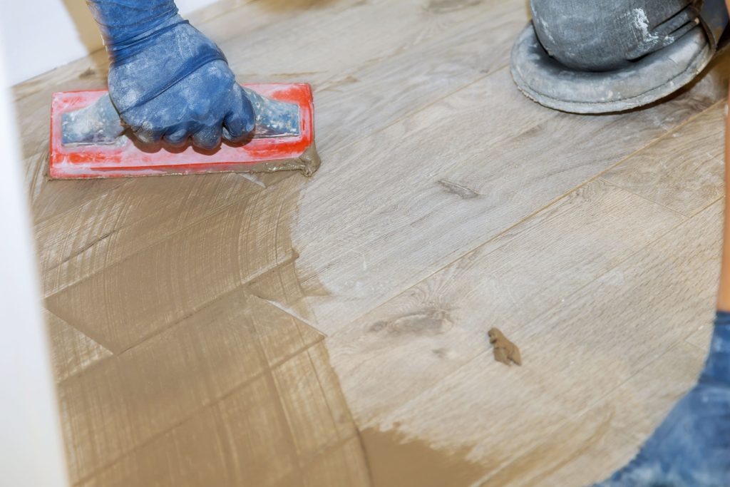 A worker in blue gloves sands a wooden floor with a red hand sander while kneeling nearby, leaving a lighter strip amid dust and scratches.