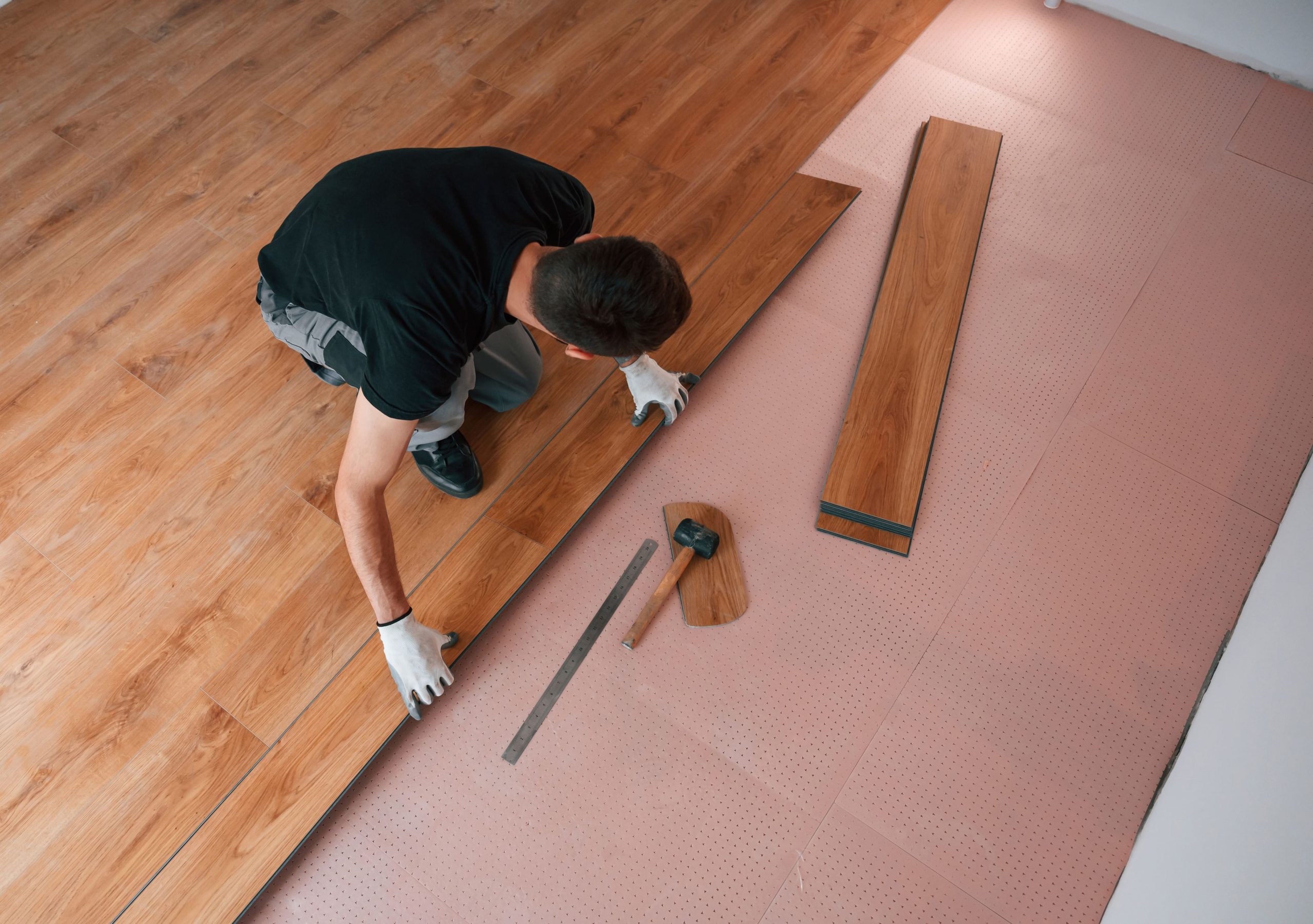 A-worker-in-gloves-kneels-while-installing-wooden-flooring-over-pink-underlayment-with-a-ruler-mallet-and-plank-nearby-scaled