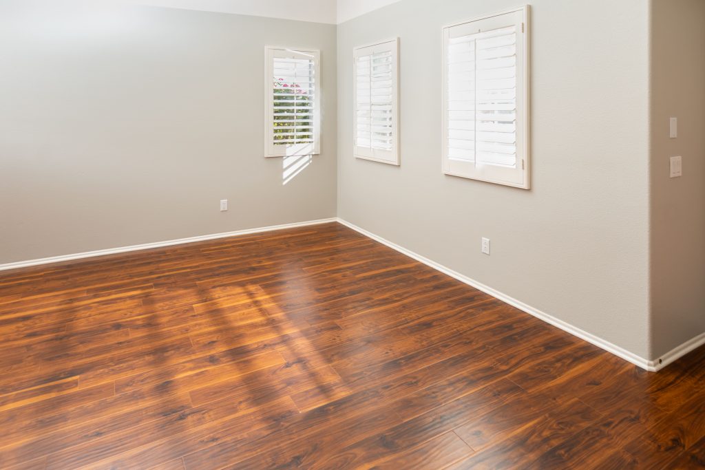 Empty room with gray walls, polished dark hardwood floors, three windows with white shutters, and electrical outlets.