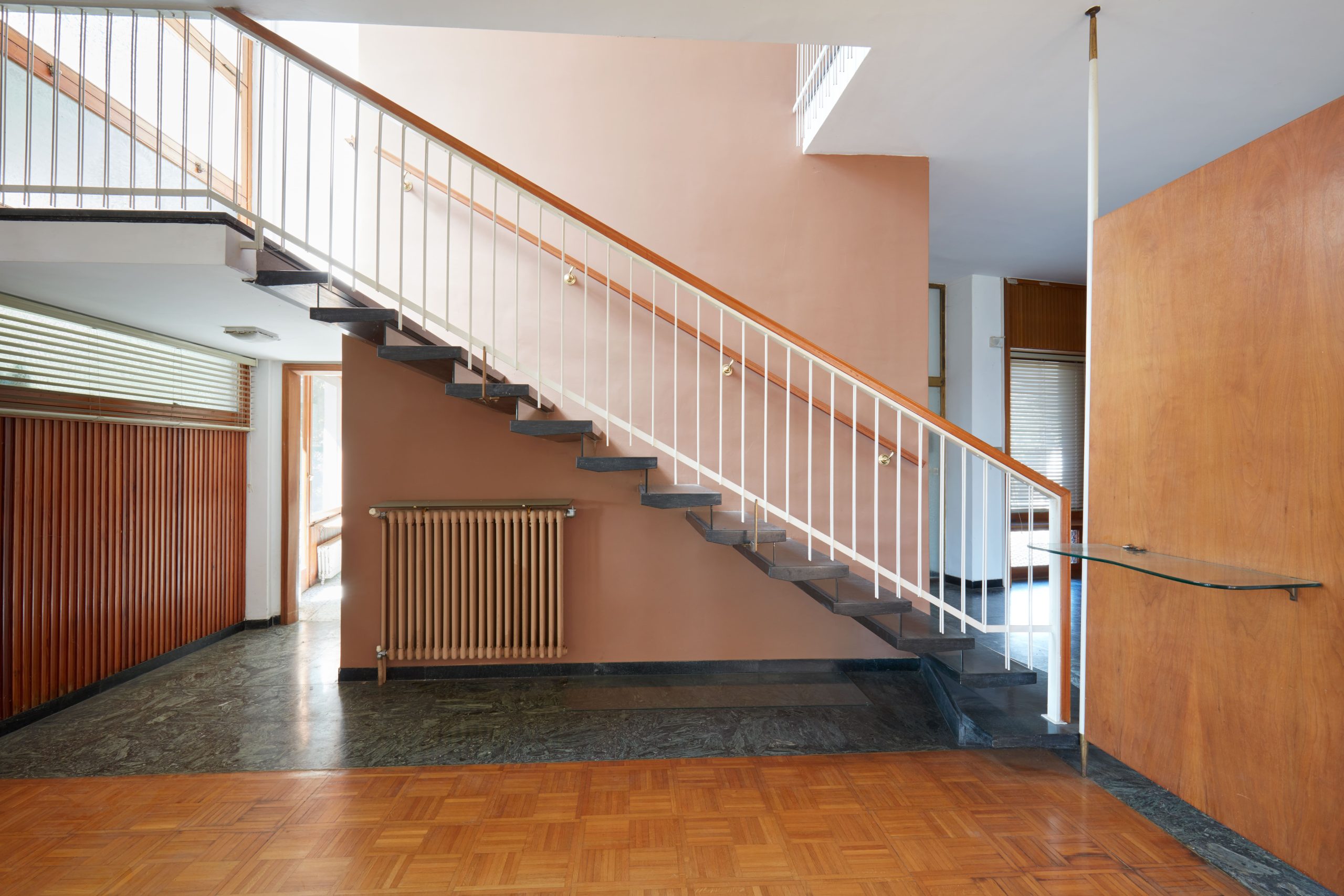 Interior with a dark stone staircase and white railings, a radiator beneath it, wood-paneled walls, windows, and parquet flooring.