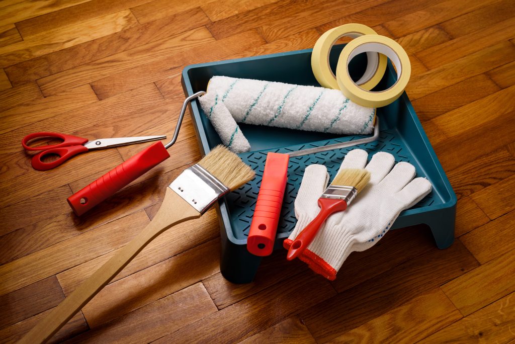Paint supplies arranged on a wooden floor, including a paint tray, roller, brushes, masking tape, scissors, and work gloves.