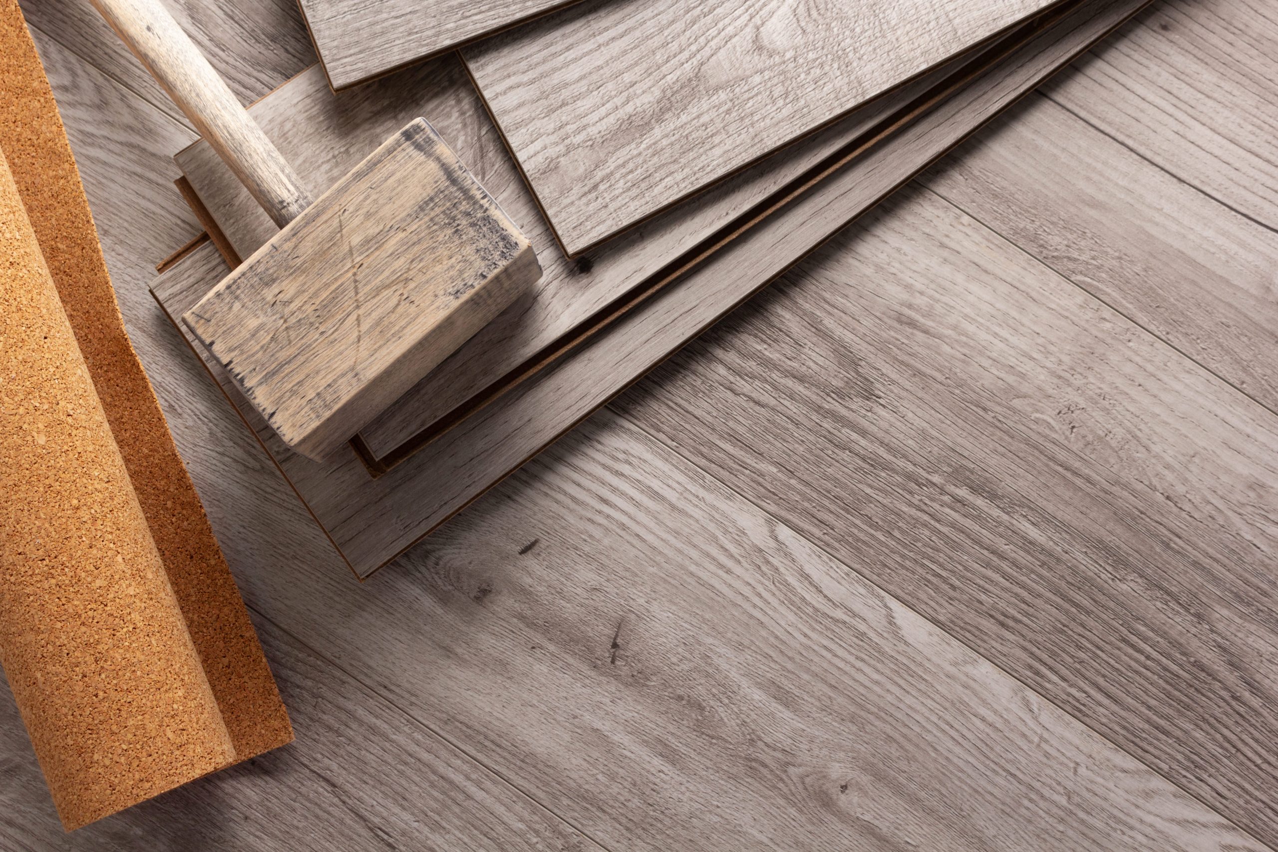 Stacked-gray-wood-flooring-planks-and-sample-pieces-lie-on-a-wood-grain-floor-beside-a-strip-of-cork-flooring