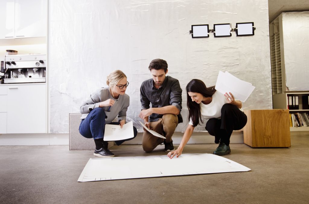 Three people sit on the floor reviewing papers and pointing at a large architectural plan in a bright room.