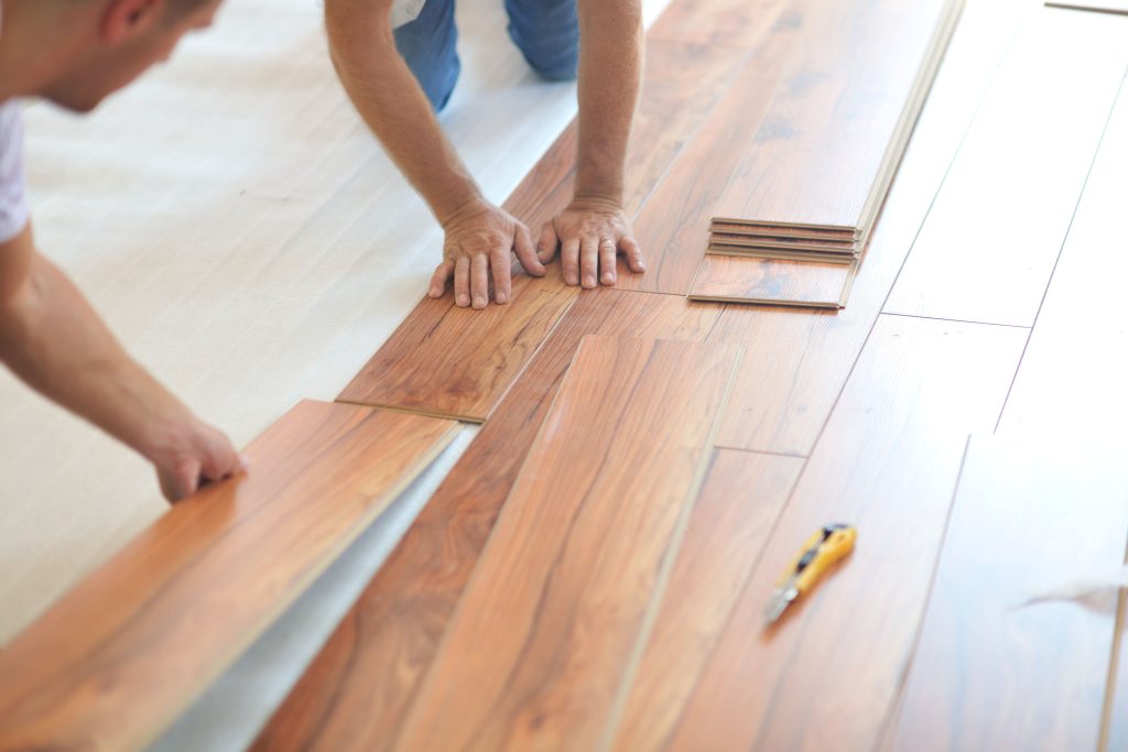 Two people install wood-look flooring, fitting planks together beside stacked flooring samples and a yellow utility knife.